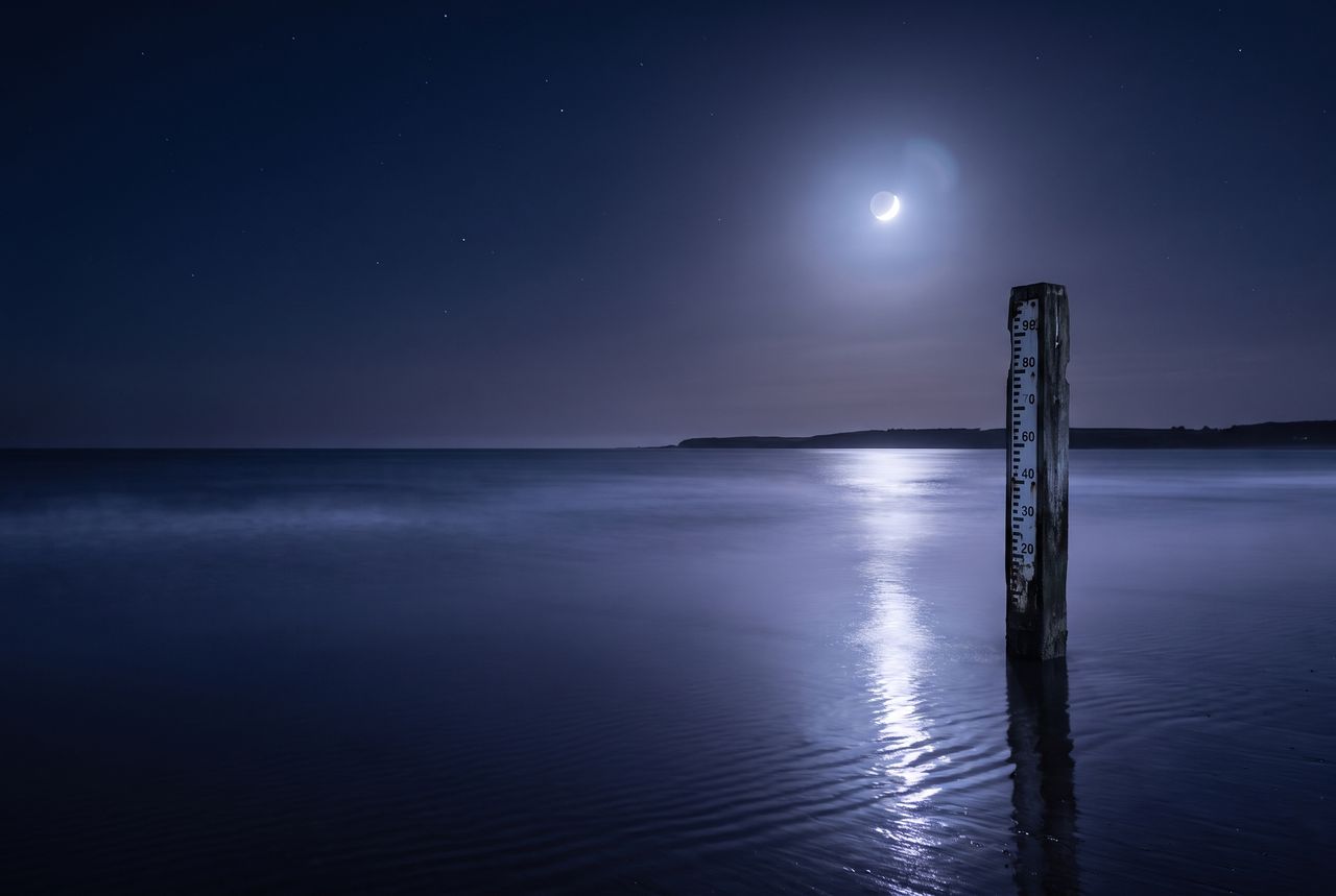 A wooden tide gauge standing in a calm night sea under a crescent moon